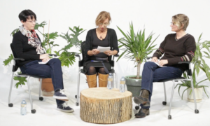 Image of three women sitting in a white room filled with plants. The middle woman is reading from a piece of paper